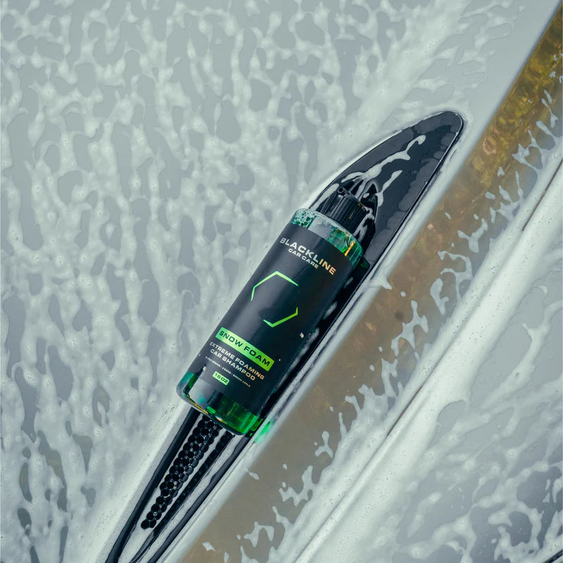 Blackline Extreme Snow Foam car shampoo bottle lying on a car surface with thick white foam, showcasing its cleaning power.