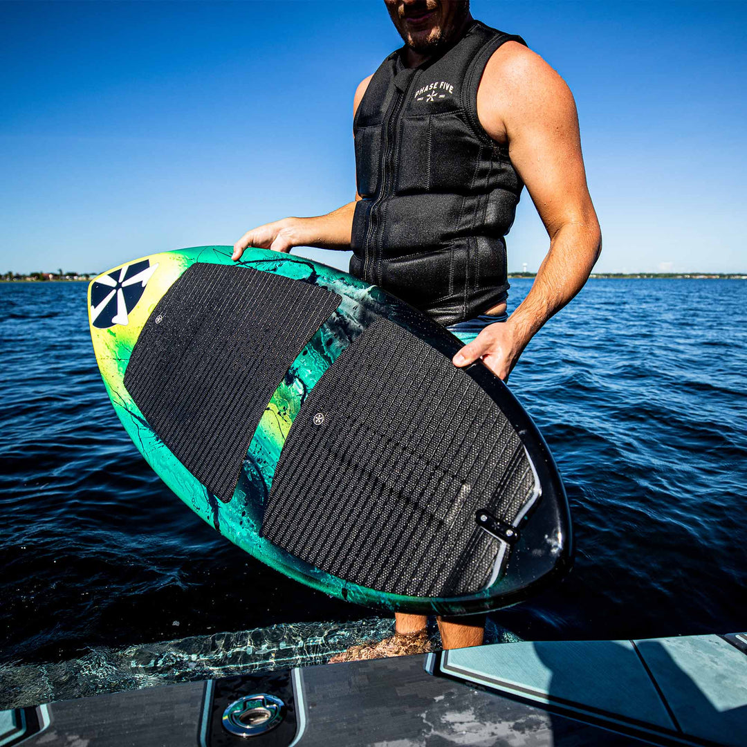 Person holding a 2025 Phase 5 Prop Skim Board on a boat, ready for water surfing, with a clear blue sky and water background.