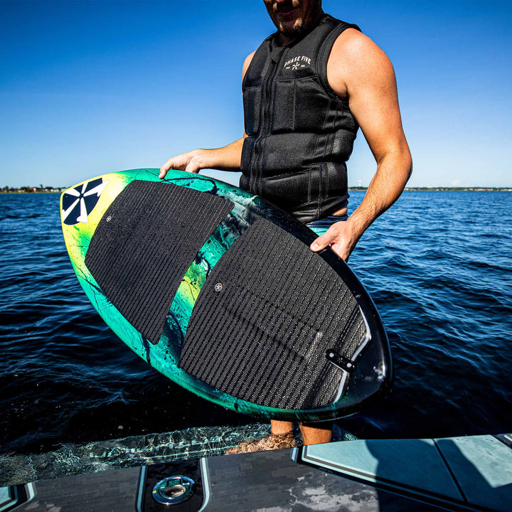 Person holding a 2025 Phase 5 Prop Skim Board on a boat, ready for water surfing, with a clear blue sky and water background.