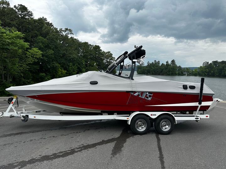 A red and white AXIS boat on a trailer, covered with a white protective cover, parked near a lake with trees and cloudy sky.