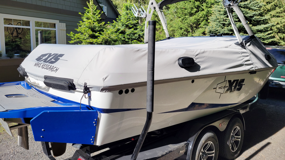 Axis A20 FatAX Tower boat on a trailer, with a white cover, black logo, and blue accents, parked outdoors near trees.