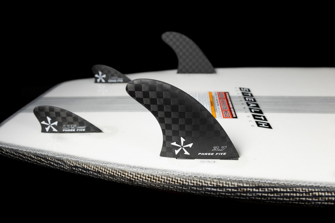 Close-up of the 2025 Phase 5 Phantom Stealth surfboard with black carbon-fiber fins, set against a black background.