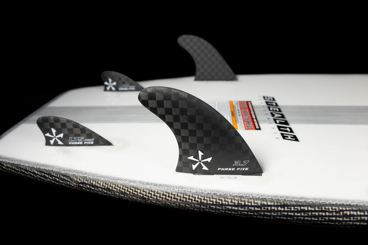 Close-up of the 2025 Phase 5 Phantom Stealth surfboard with black carbon-fiber fins, set against a black background.