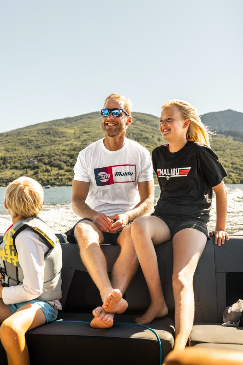 Smiling family on a boat wearing Malibu Statement Tees, enjoying sunny weather with scenic hills and water in the background.