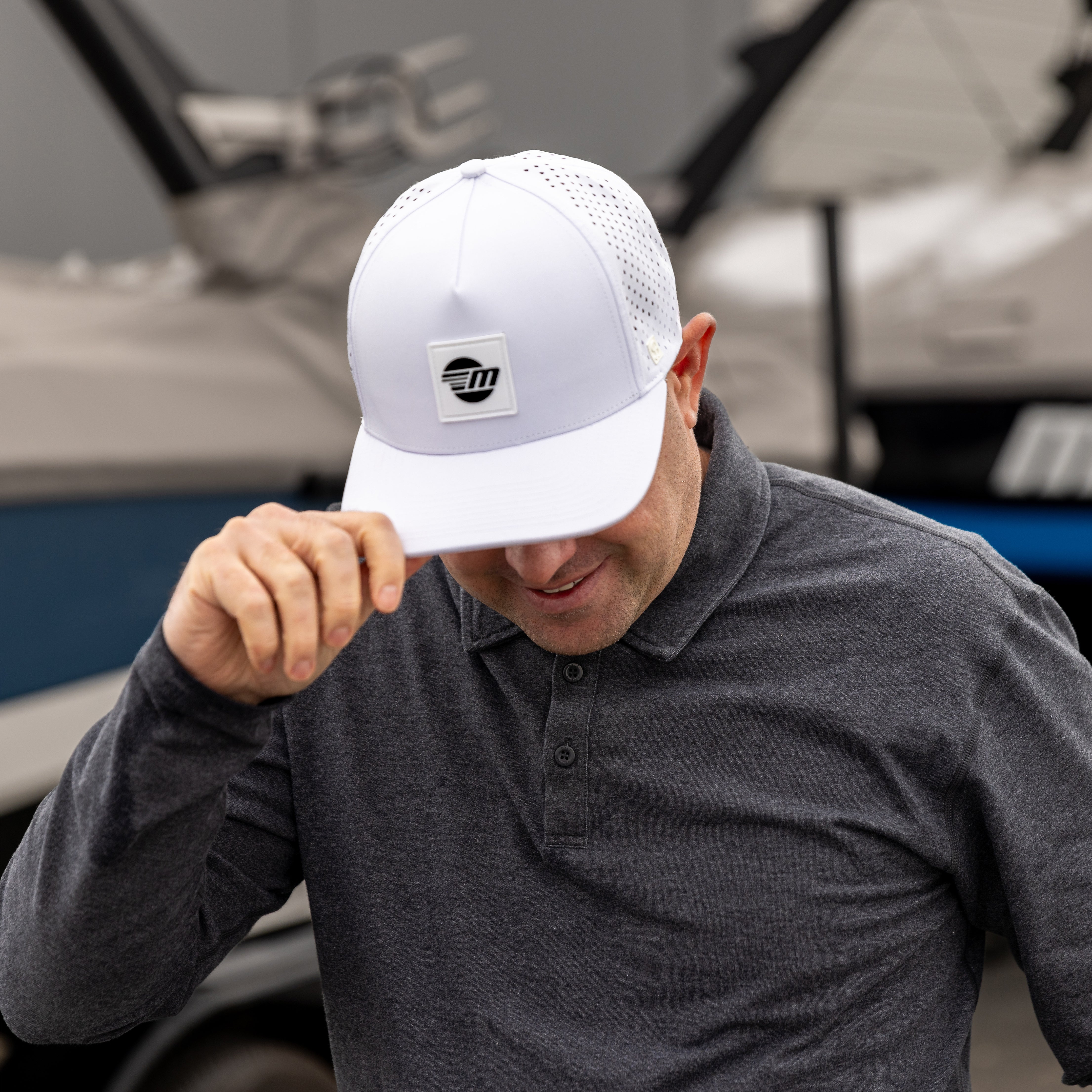 Man wearing a white Malibu Swell hat, adjusting the brim, outdoors near boats, smiling, in a gray collared shirt.