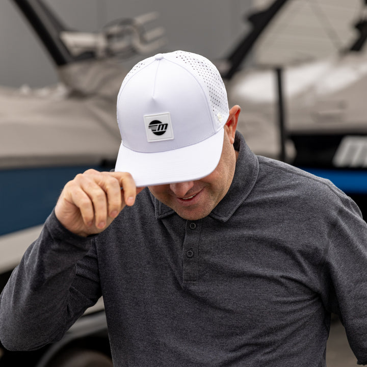 Man wearing a white Malibu Swell hat, adjusting the brim, outdoors near boats, smiling, in a gray collared shirt.