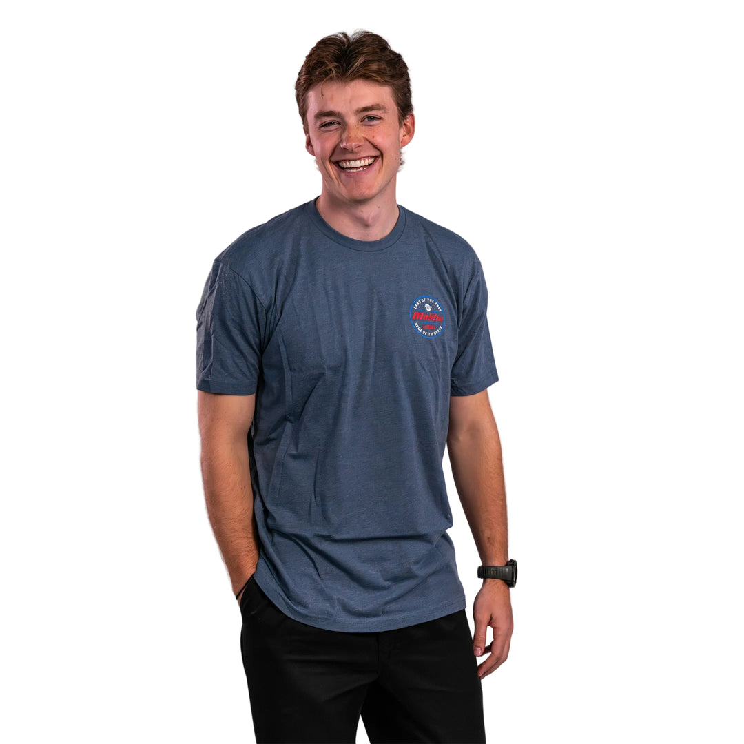 A young man wearing a blue Malibu Brave Tee, smiling confidently with hands in pockets, showcasing a casual style.