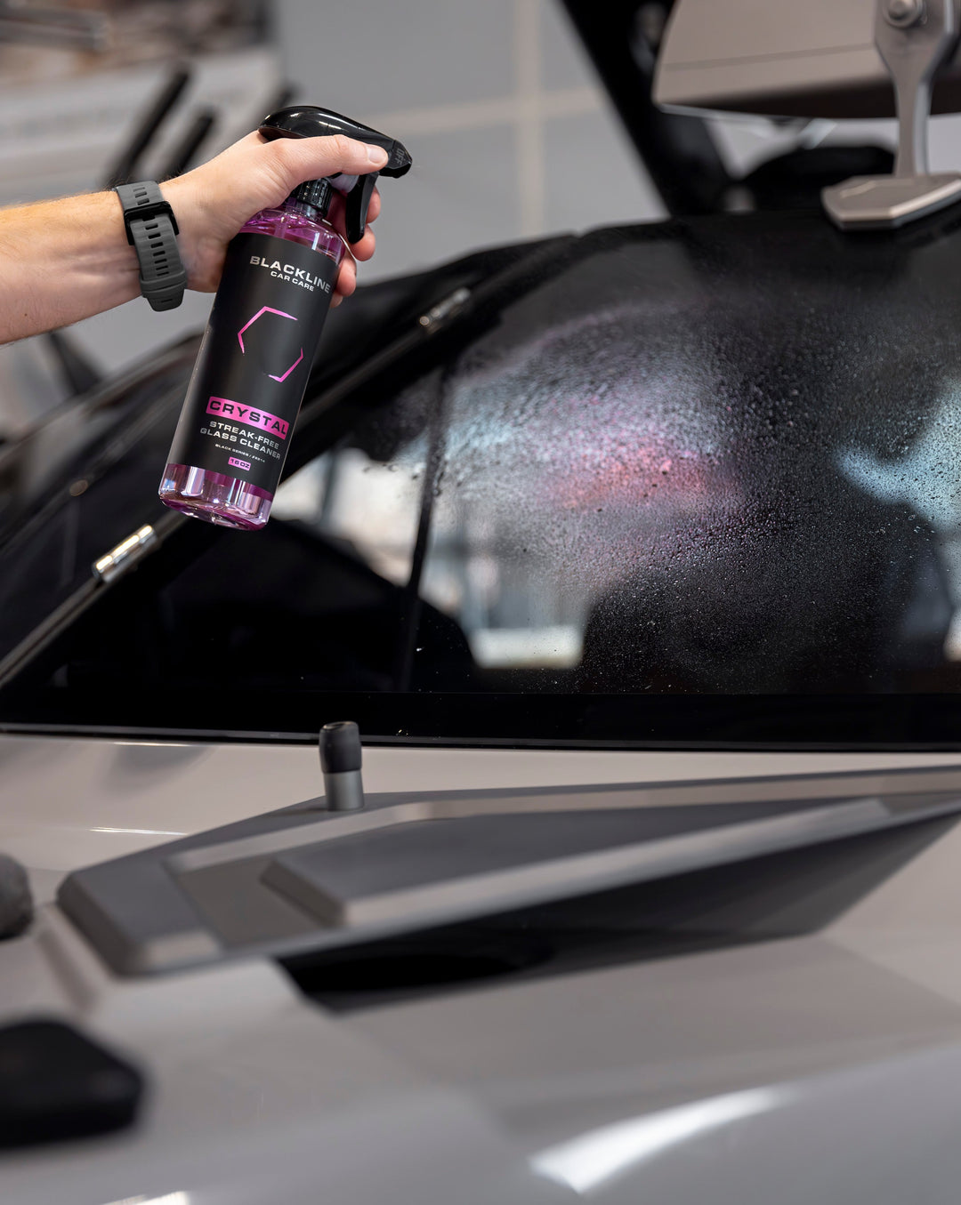 Blackline Crystal Glass Cleaner being sprayed on a car windshield to remove streaks and dirt for a clear view.