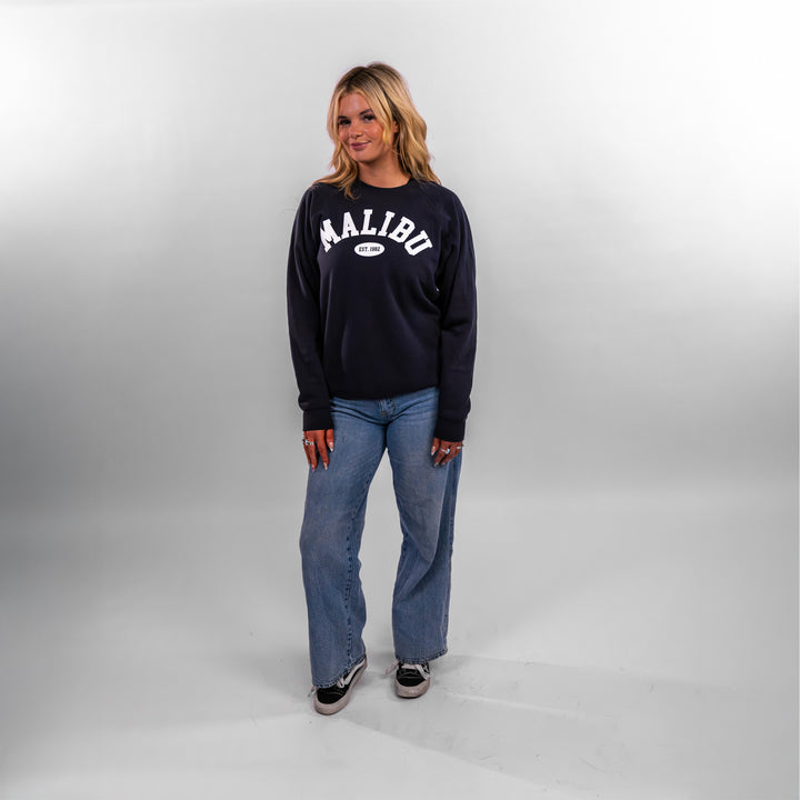Model wearing a navy Malibu Varsity Crewneck sweatshirt with the word "MALIBU" printed across the front, paired with light blue jeans.