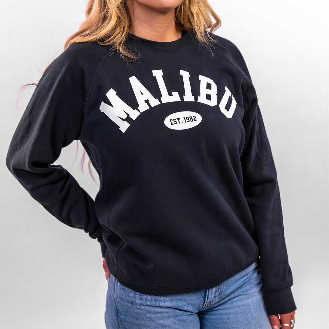 Woman wearing a black Malibu Varsity Crewneck sweatshirt featuring the word "MALIBU" and established year 1982.