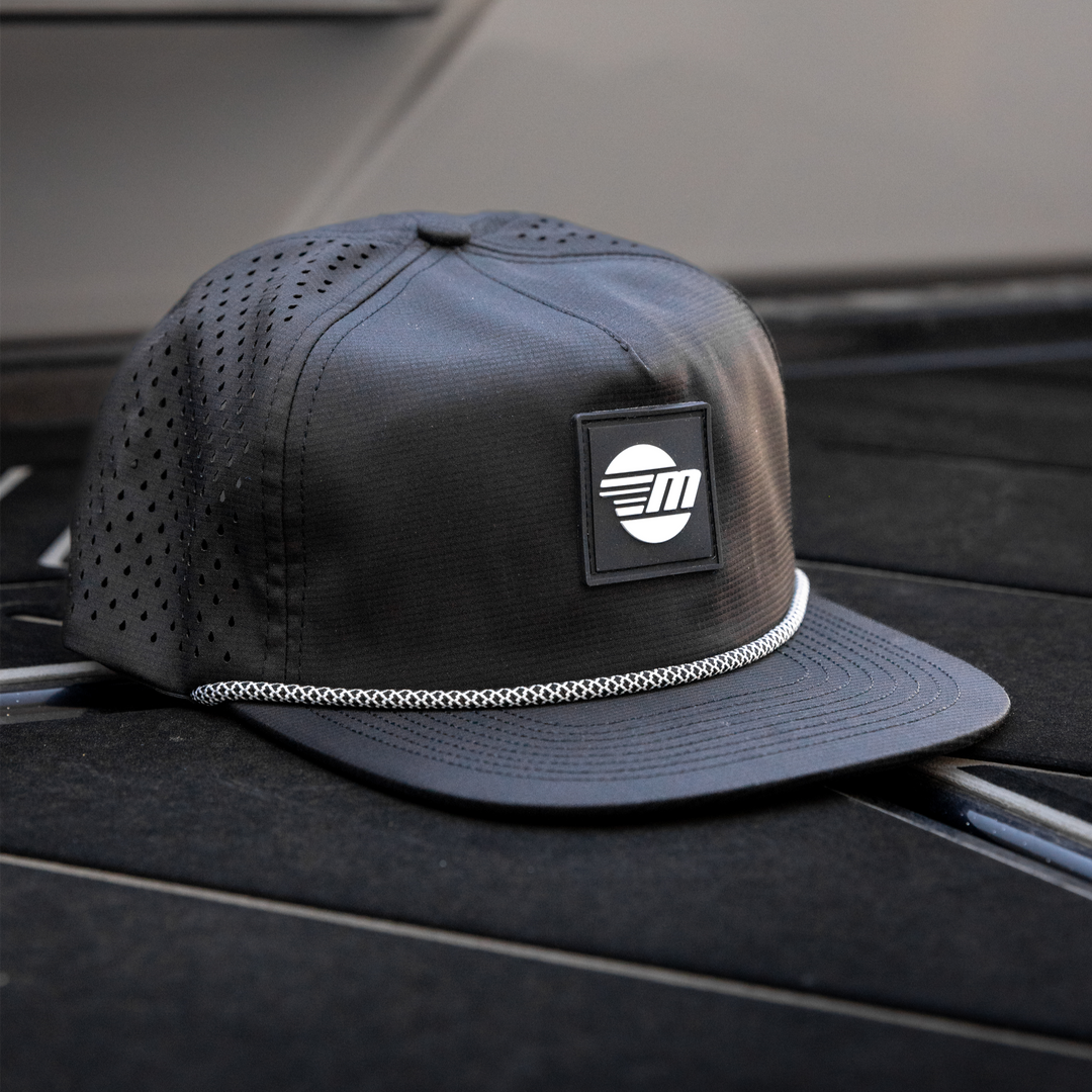 Black Malibu Dockside Hat with a logo patch, mesh panels, and a white braided trim, placed on a boat deck.