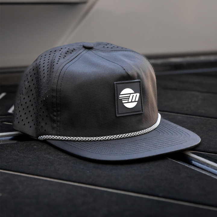 Black Malibu Dockside Hat with a logo patch, mesh panels, and a white braided trim, placed on a boat deck.