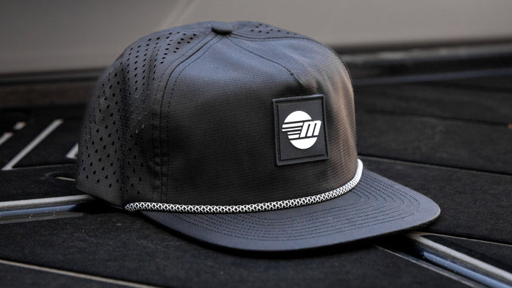 Black Malibu Dockside Hat with a mesh crown, logo patch, and white braided trim, placed on a dark surface.