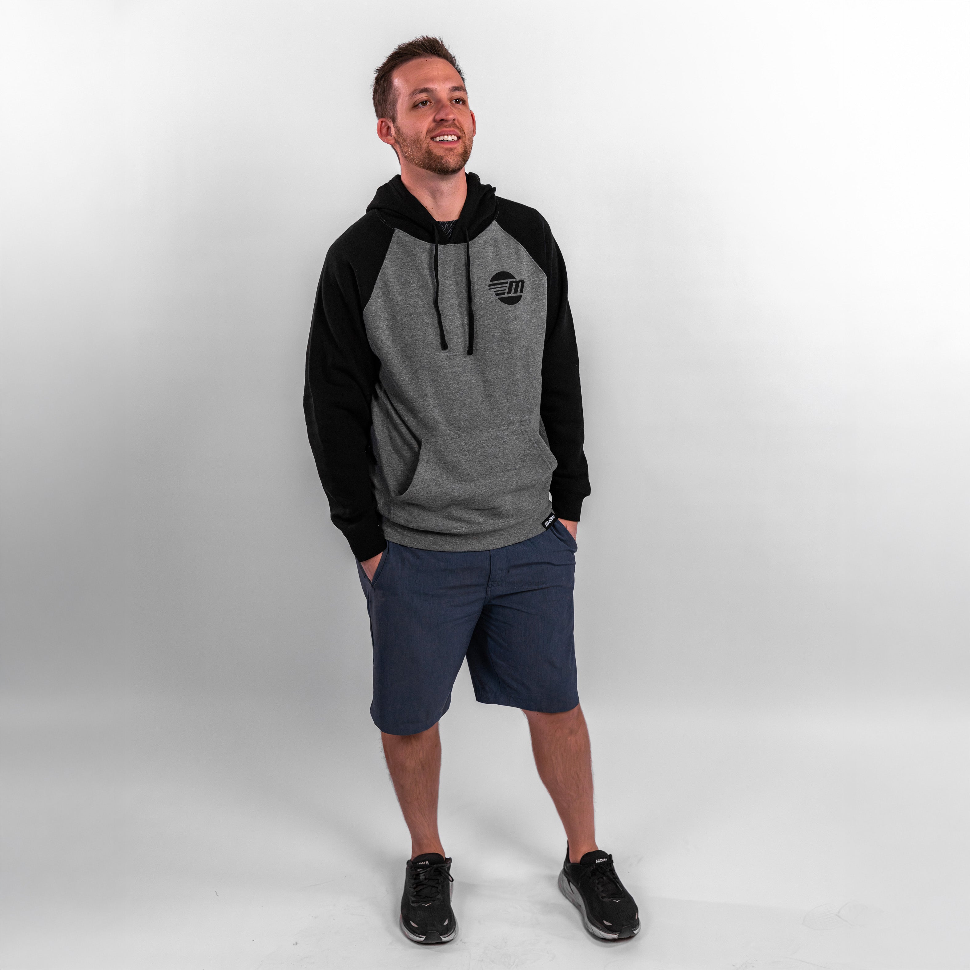 A man stands smiling in a gray and black Malibu hoodie, paired with shorts and sneakers, showcasing a casual look.