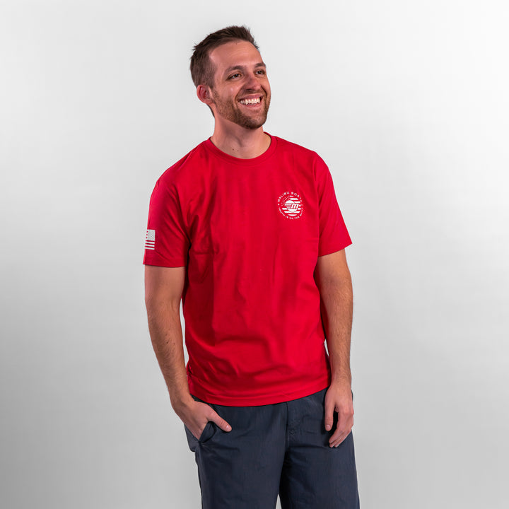Man wearing a red Patriot Tee with a logo on the front and an American flag on the sleeve, smiling against a neutral background.