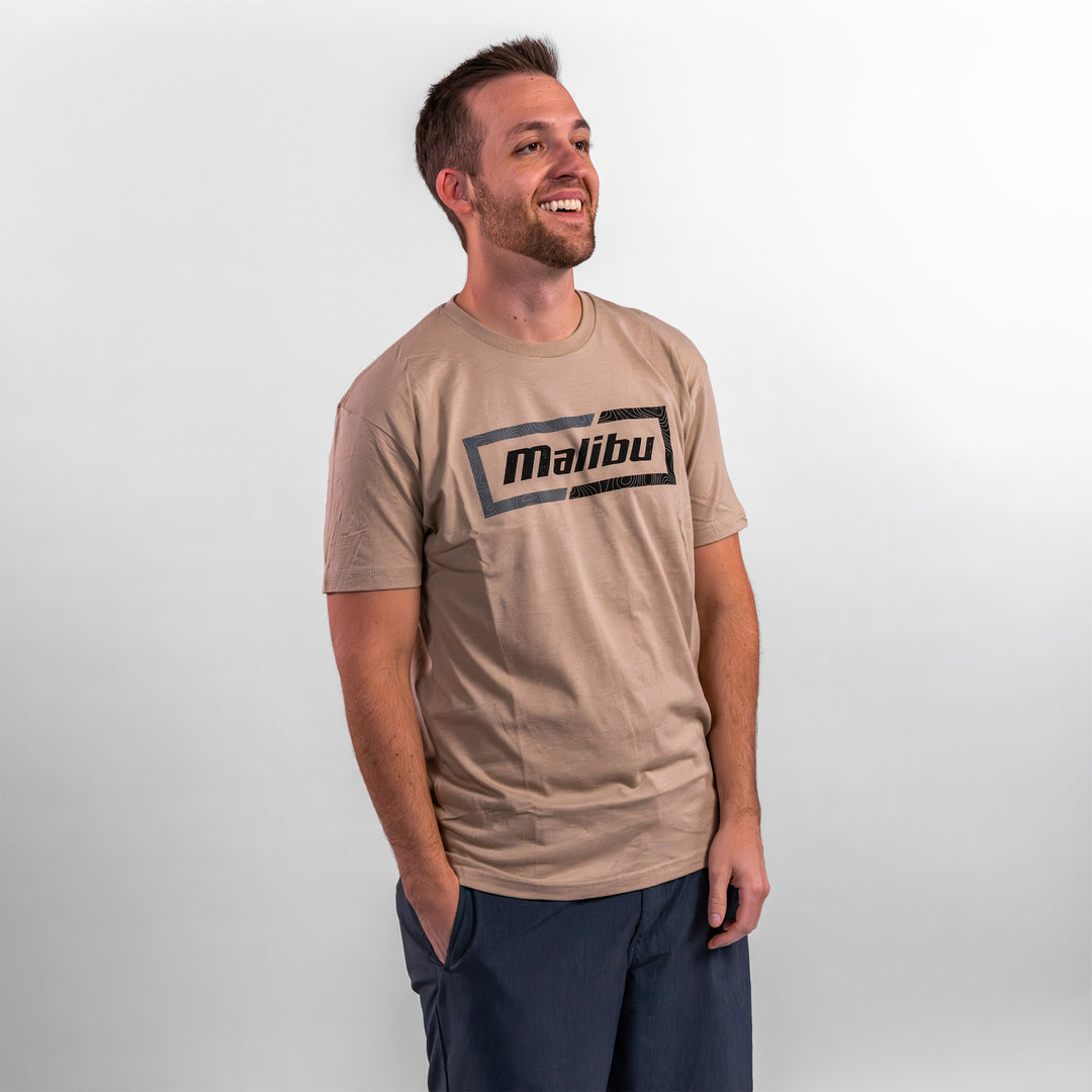 Man wearing a beige Malibu Topo Tee featuring a bold black logo, smiling against a neutral background.