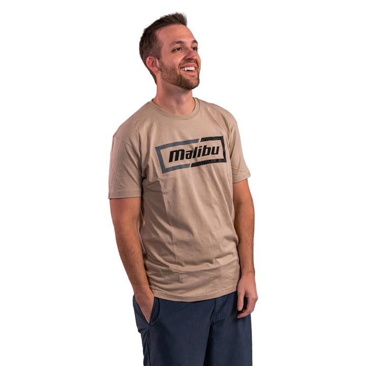 Man wearing a beige Malibu Topo Tee featuring a bold black logo, smiling against a neutral background.