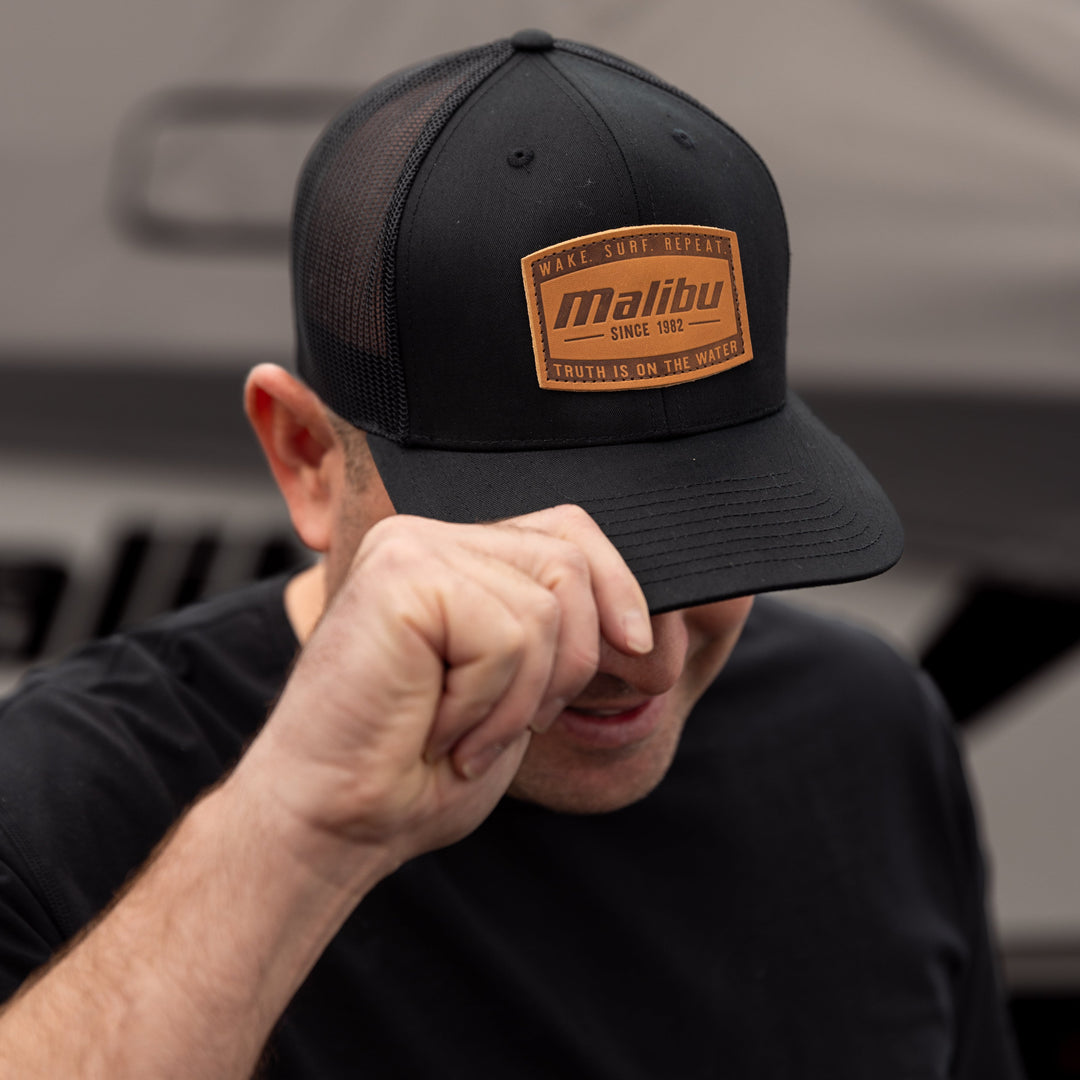 A man wearing a black trucker-style ranger hat with a brown patch that says "Malibu" and "Since 1982," adjusting the brim with his hand.