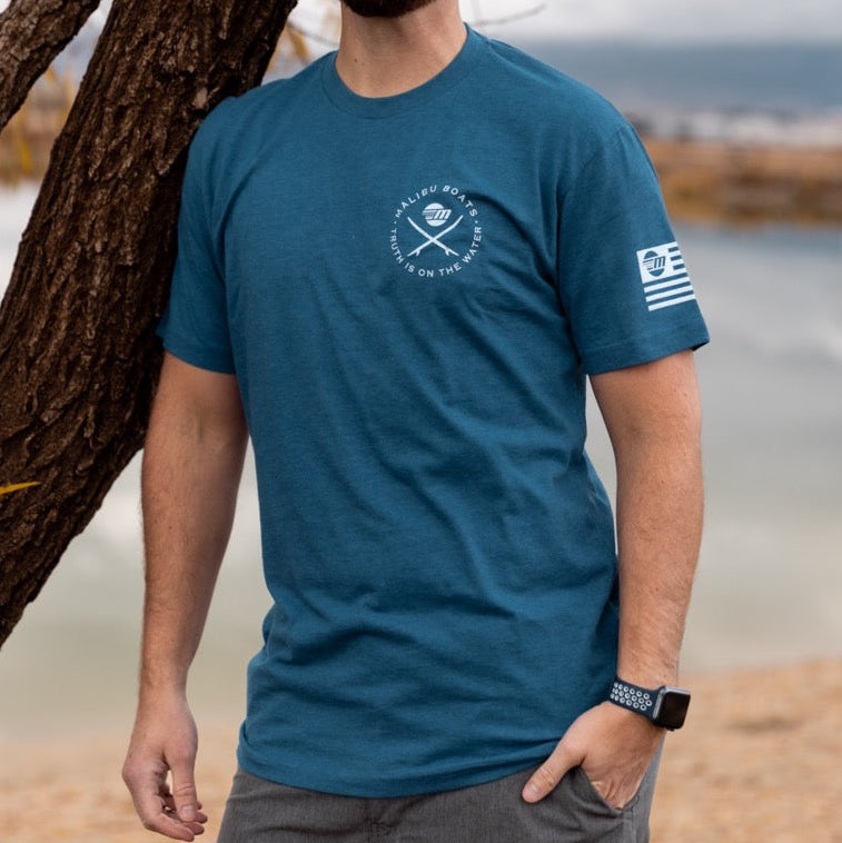 Man wearing a blue Malibu Endless Wake Tee (Tall) with boat logo and text, standing outdoors by water and a tree.