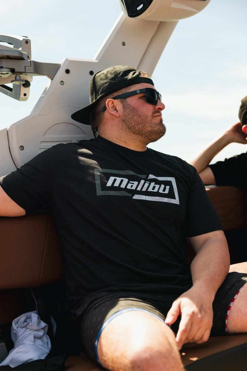 Man wearing a black Malibu Topo Tee, sunglasses, and a backward cap, relaxing on a boat with a telescope in the background.
