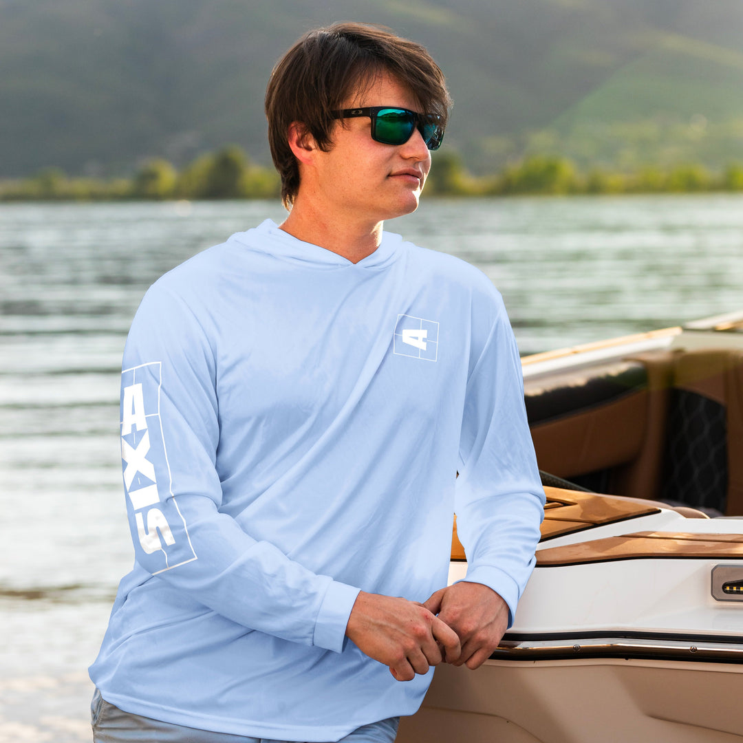 Man wearing a light blue AXIS shaded shredder sun hoodie with logo, standing by a boat on a lake with mountains in the background.