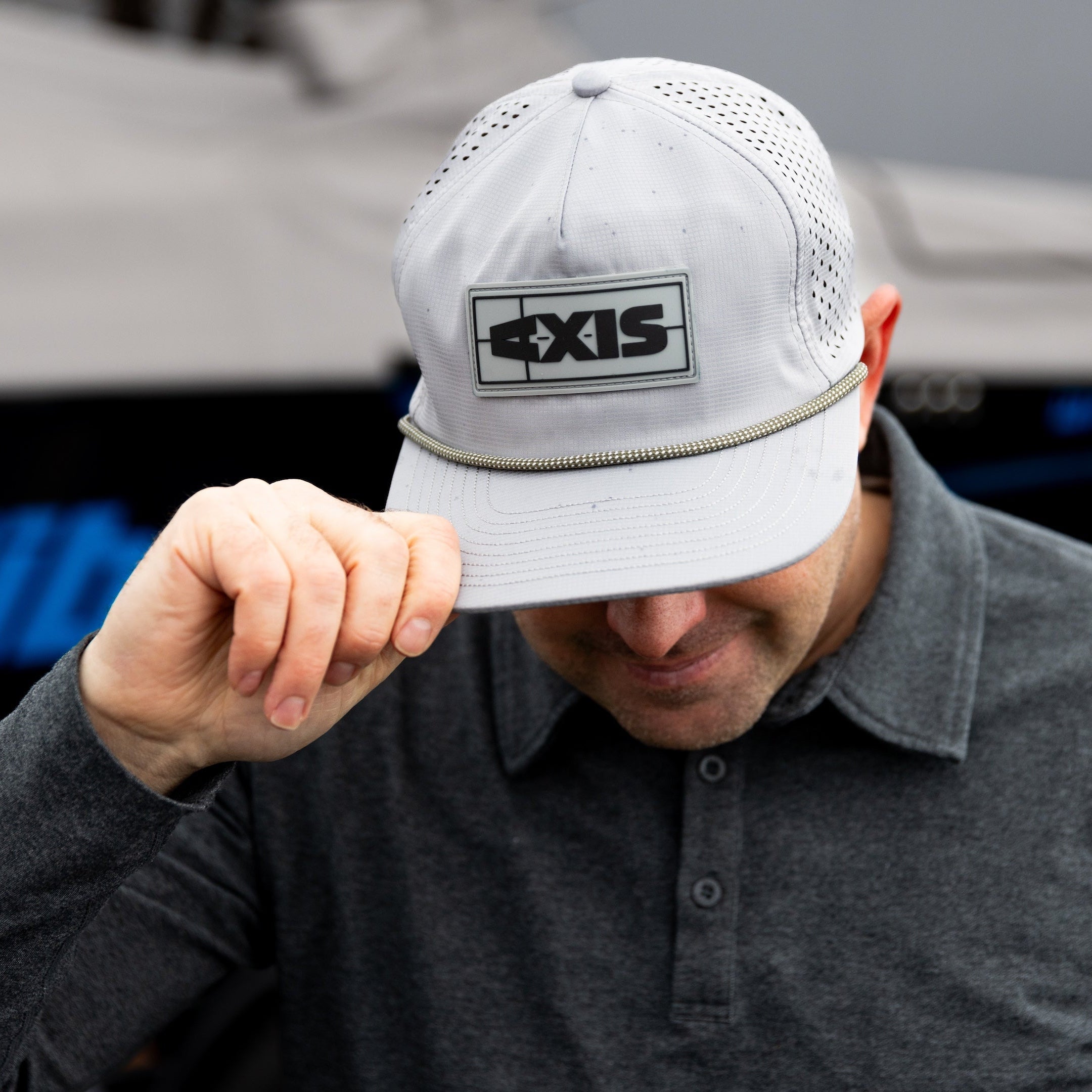 A man wearing a white Axis Dockside Hat with a logo patch and decorative rope, adjusting the brim in front of a boat.
