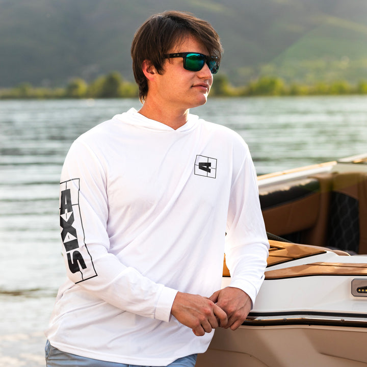 Man wearing a white Shaded Shredder Sun Hoodie by AXIS with black logo details, standing near a boat on a lake.