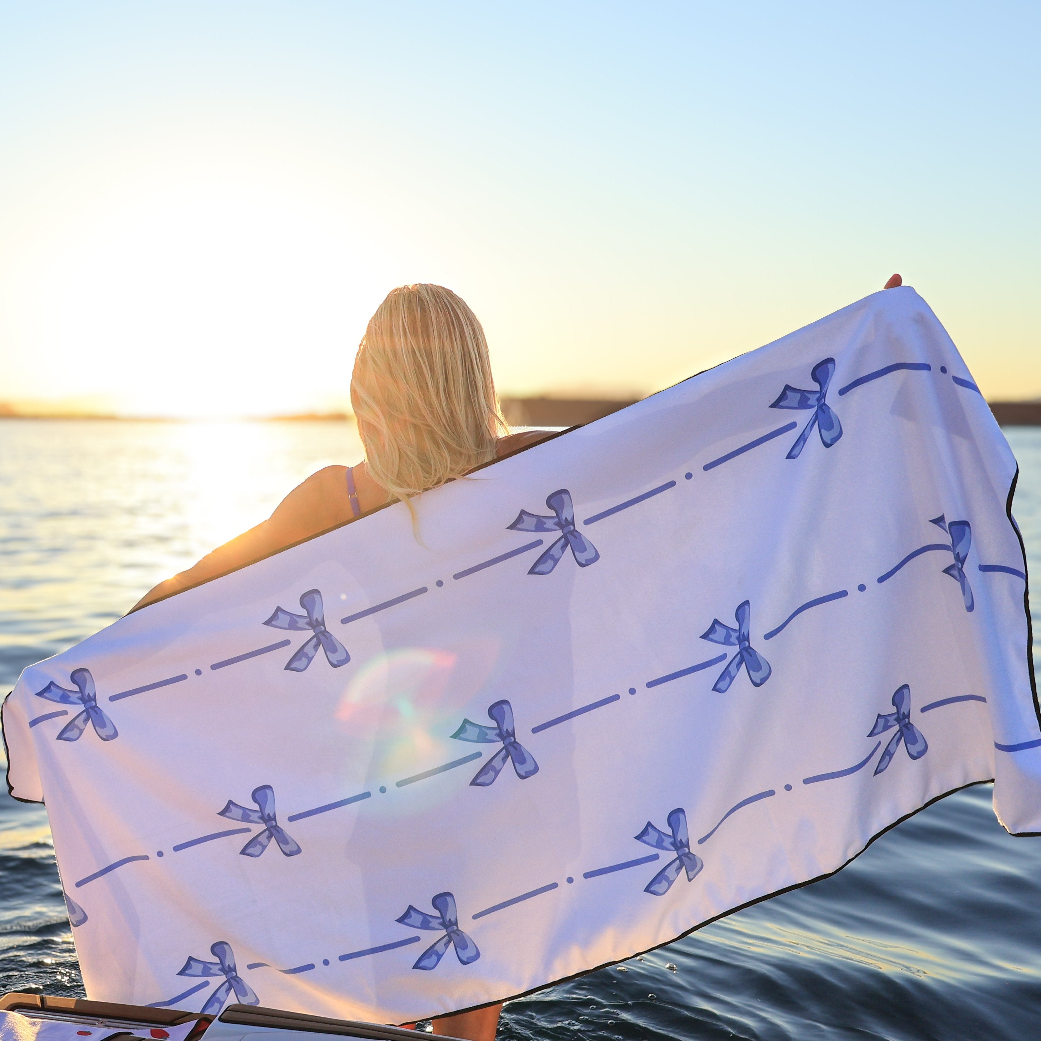 Woman holding a blue bow-patterned lake towel by the water at sunset, with the sun low on the horizon.