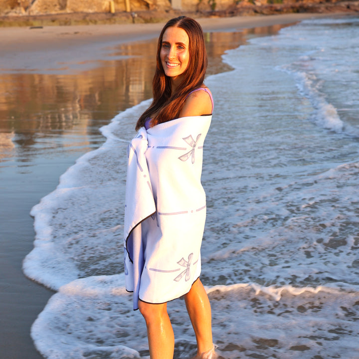 Woman standing on the beach wrapped in a white and blue Wake Head Blue Bliss Lake Towel, with waves and shoreline in the background.