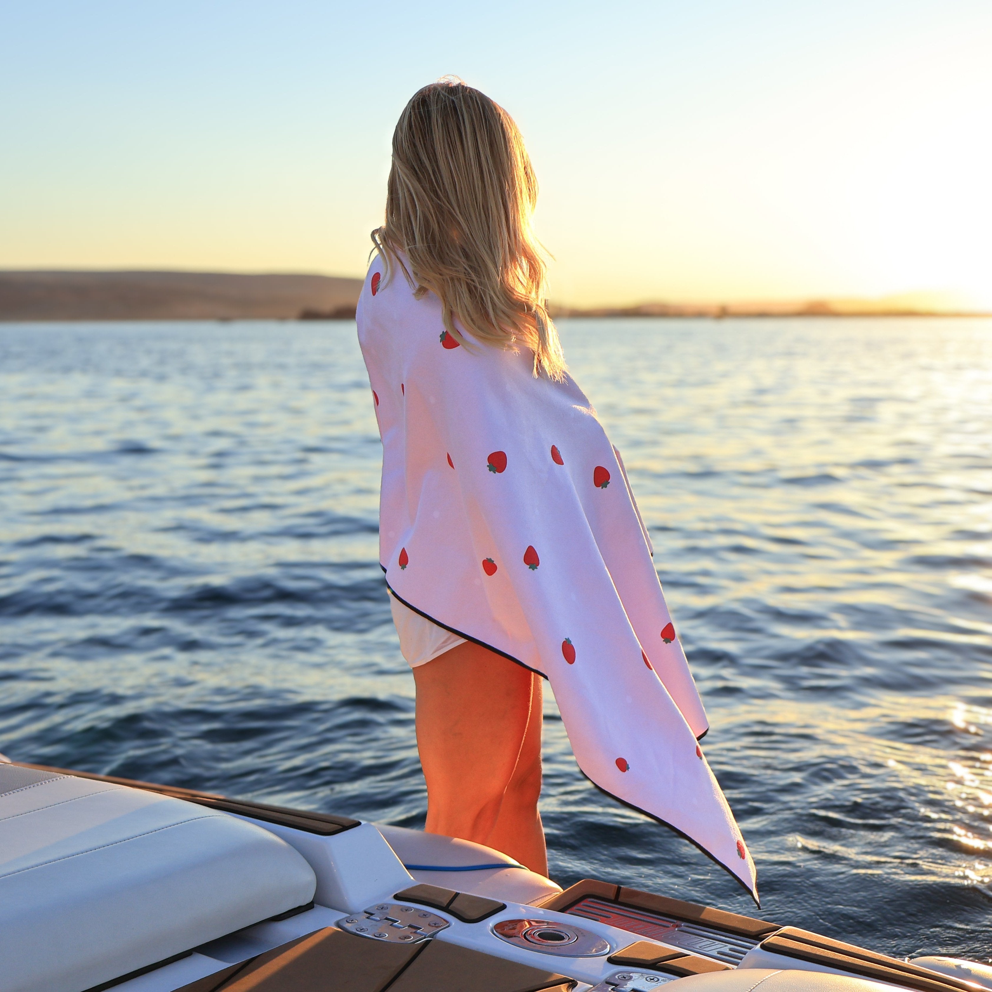 Woman on boat with a white towel featuring red strawberries, enjoying sunset by the water.