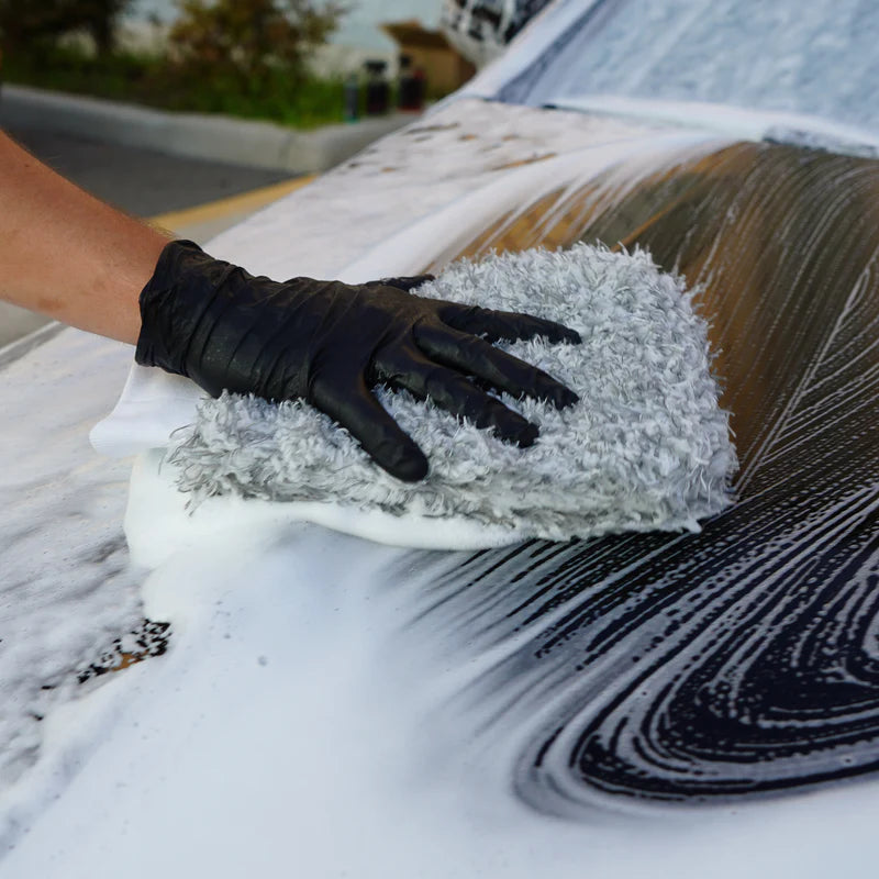 A person wearing a black glove uses a microfiber wash mitt to clean soapy water off a car&