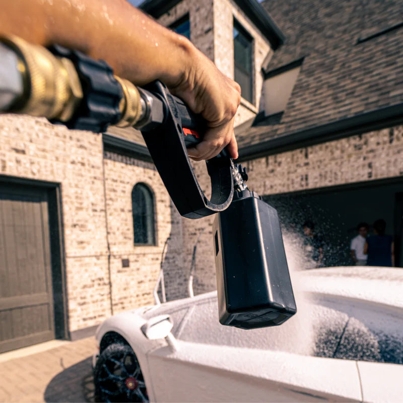 Person using a foam cannon to spray Blackline Extreme Snow Foam on a white car outside a house.