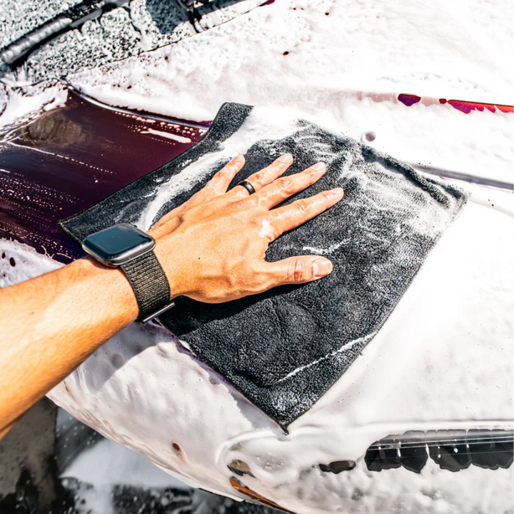 A hand using a synthetic clay towel on a soapy car surface, demonstrating effective cleaning and detailing.