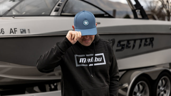 Man wearing a black Malibu hoodie and blue Malibu Truth hat, standing in front of a boat.