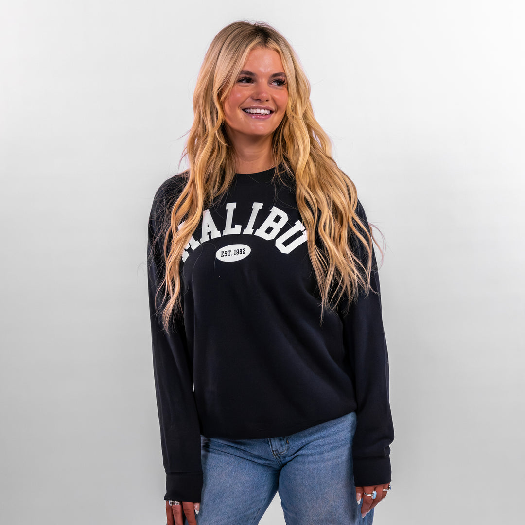 Model wearing a black Malibu Varsity Crewneck sweatshirt with white lettering, styled with light blue jeans.
