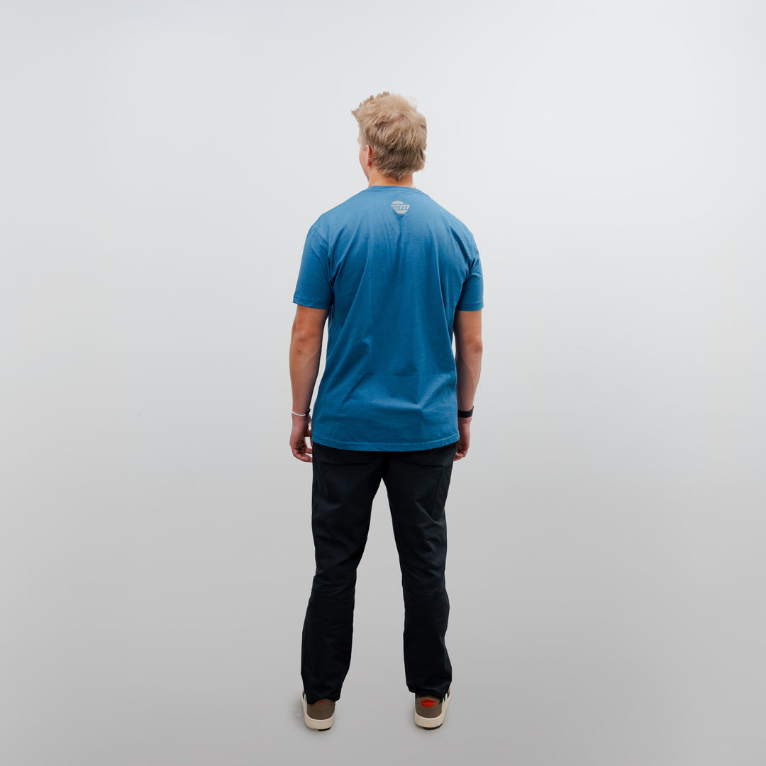Back view of a person wearing a blue Malibu Icon Tee, black pants, and beige sneakers, standing against a plain white background.