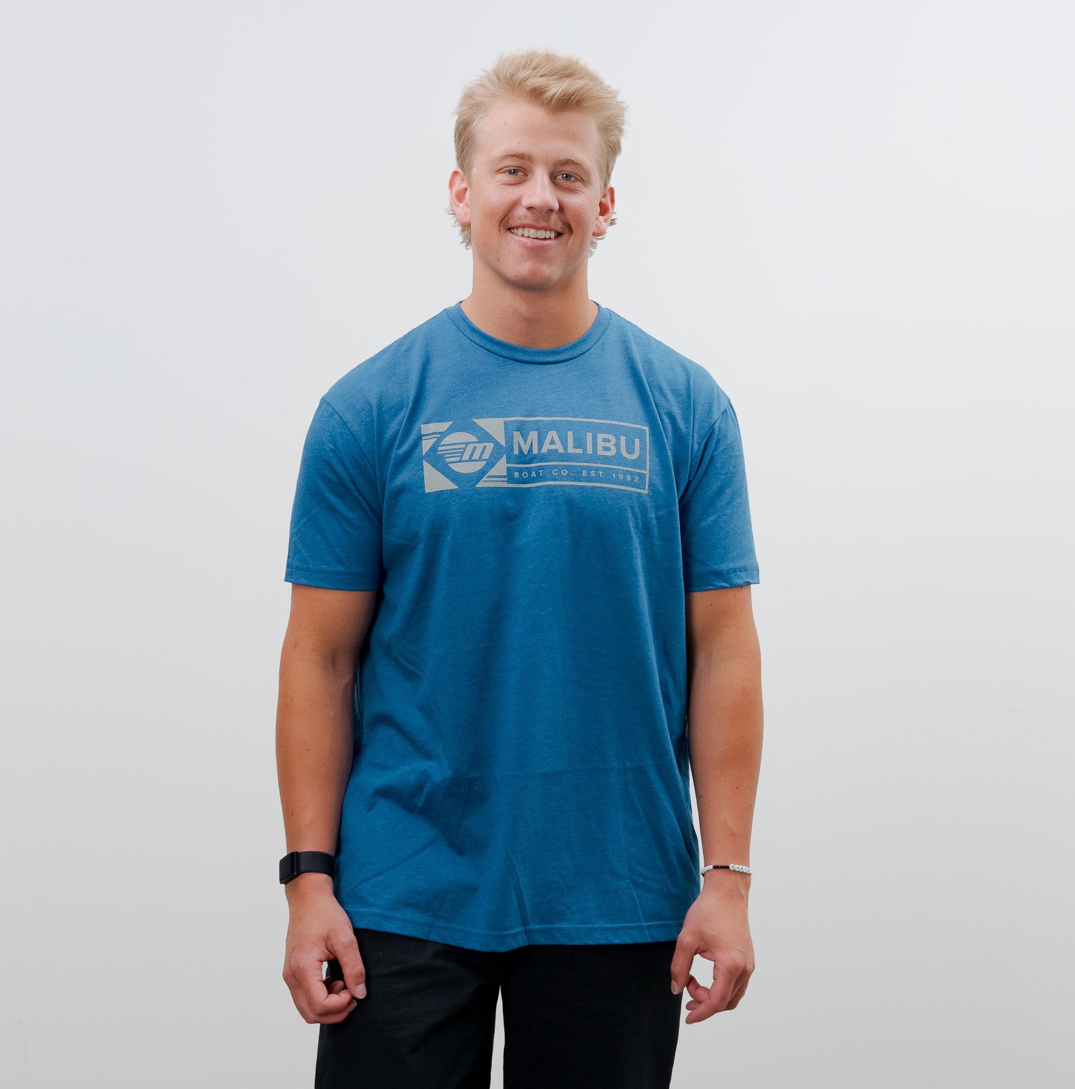 Young man wearing a blue Malibu Icon Tee, black pants, and sneakers, smiling against a plain white background.