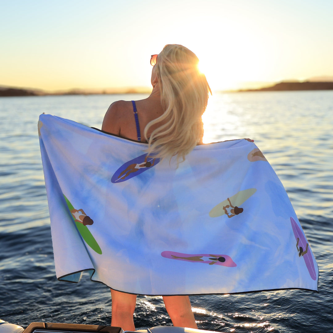 Woman holding a Wake Head Tide Chaser Lake Towel with colorful surfers design, standing by the water at sunset.