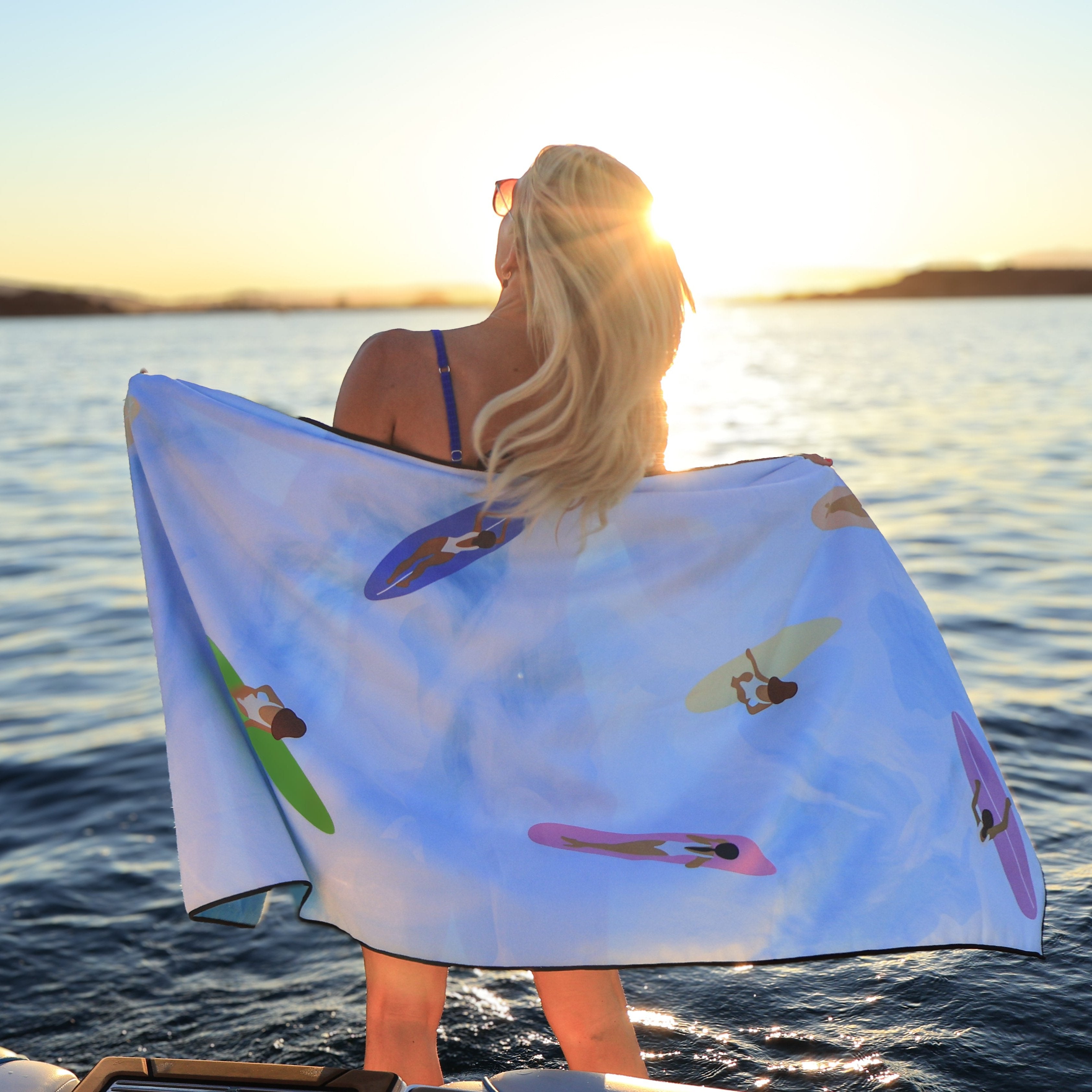 Woman holding a Wake Head Tide Chaser Lake Towel with colorful surfers design, standing by the water at sunset.