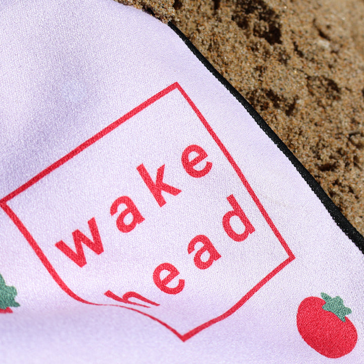 Close-up of a white Lake Towel with red "Wake Head" text and strawberry illustrations, lying on sandy beach.