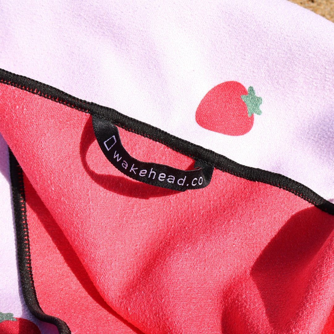 Close-up of a pink and purple berry-themed lake towel with a strawberry print and a black tag reading "Wakehead.co".