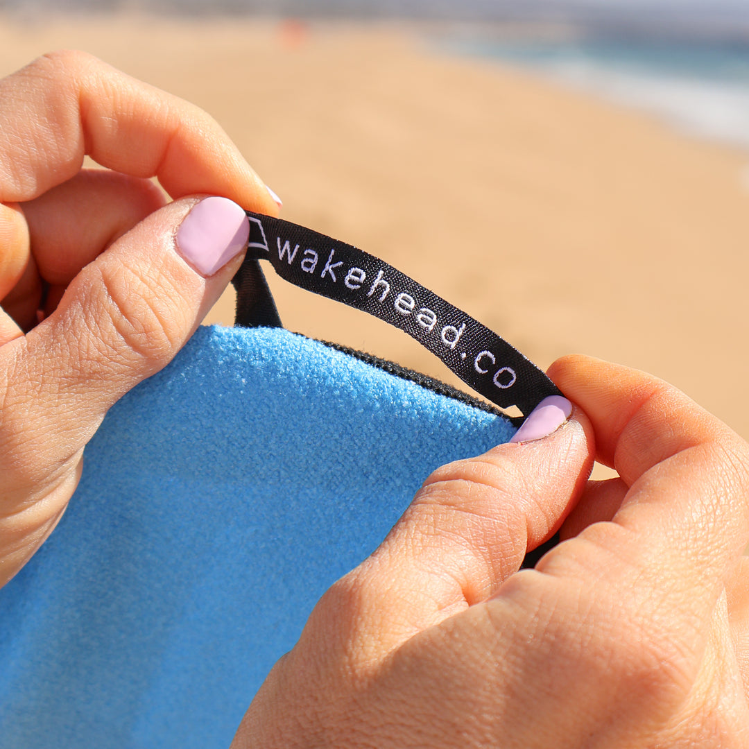 Close-up of hands holding a blue Tide Chaser Lake Towel with a black tag reading "wakehead.co" at the beach.