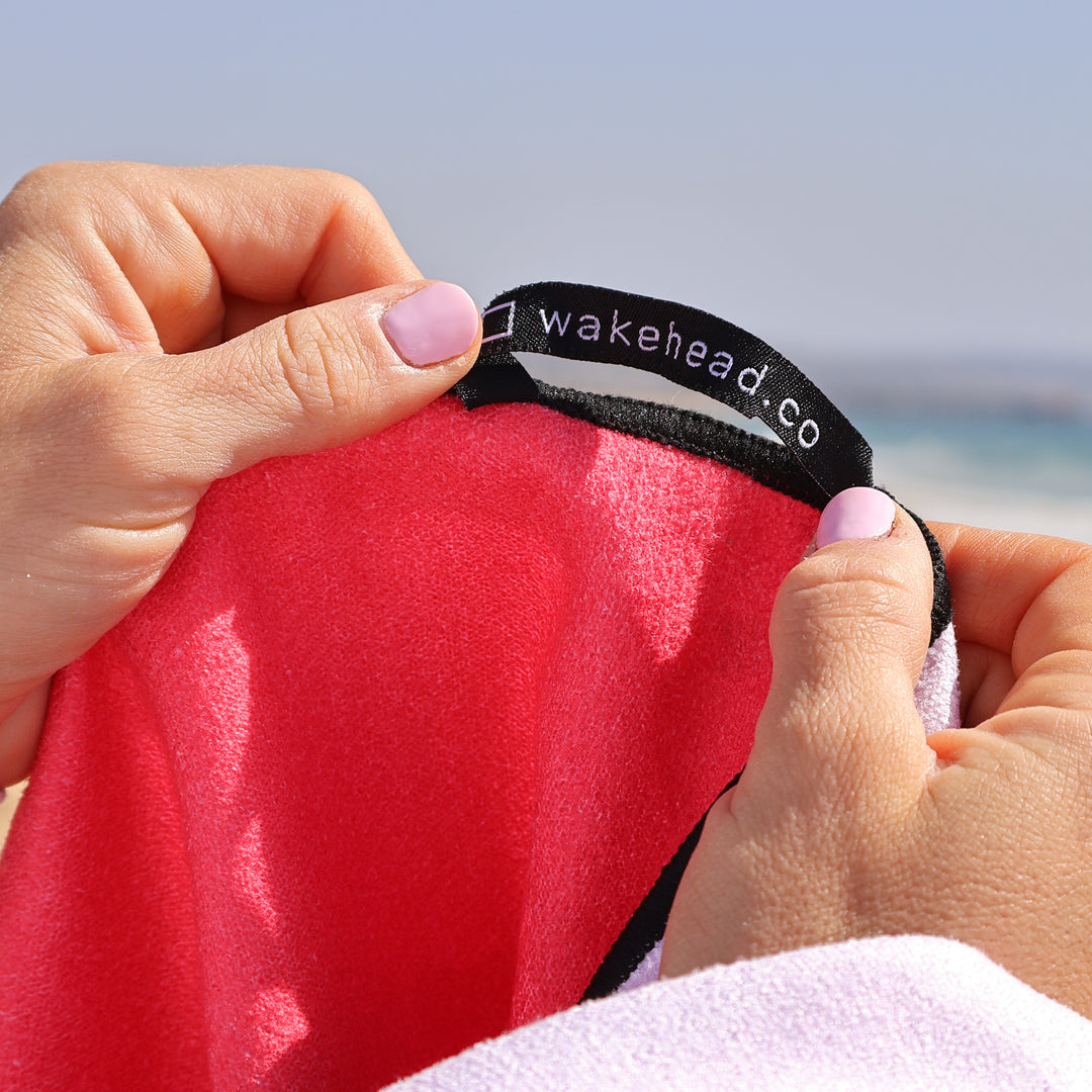 Close-up of hands holding a bright pink Lake Towel with a black tag reading "wakehead.co" against a beach background.