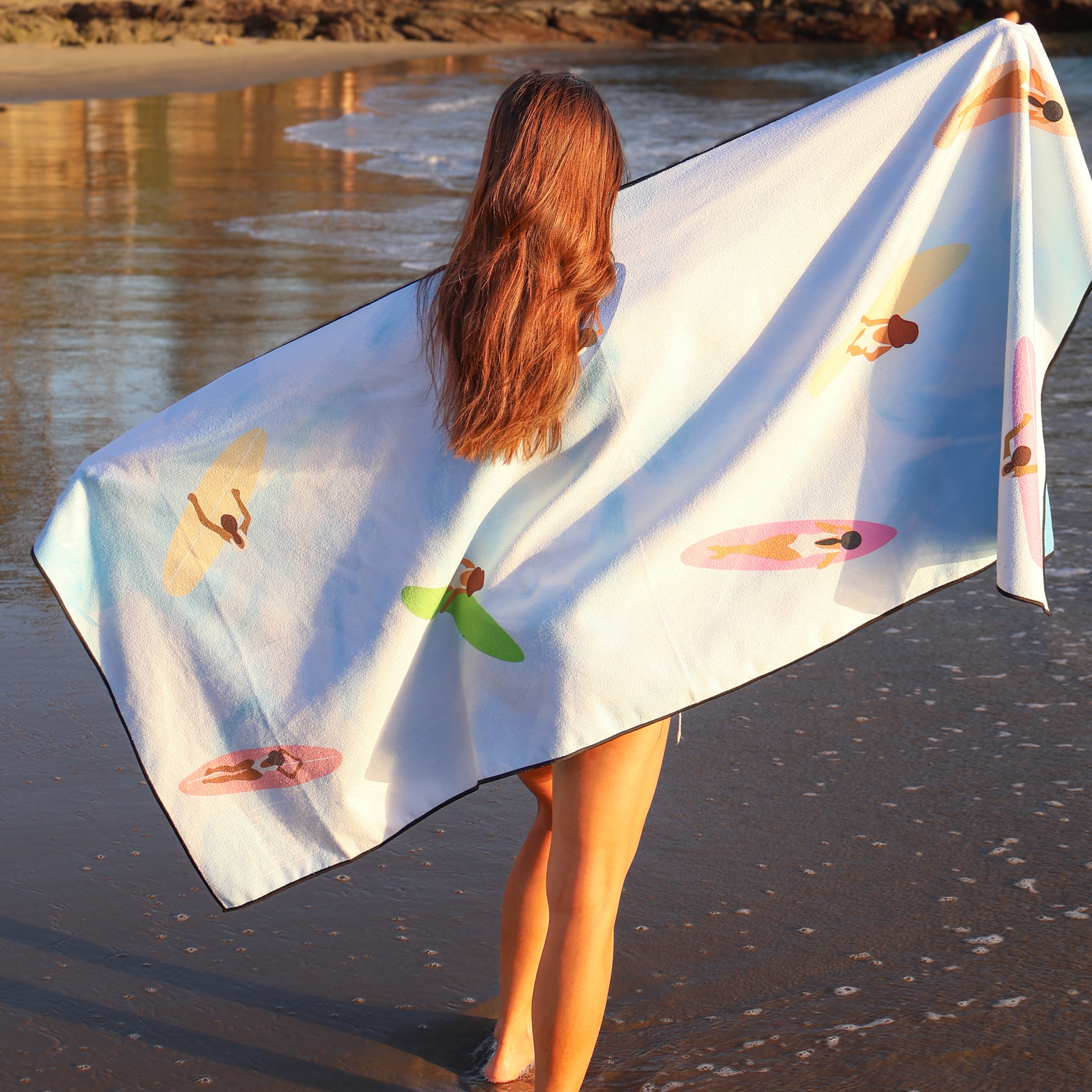 Woman with long red hair holding a white lake towel with colorful surfer girl prints, standing at the shoreline.