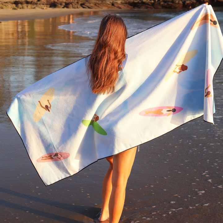 Woman with long red hair holding a white lake towel with colorful surfer girl prints, standing at the shoreline.