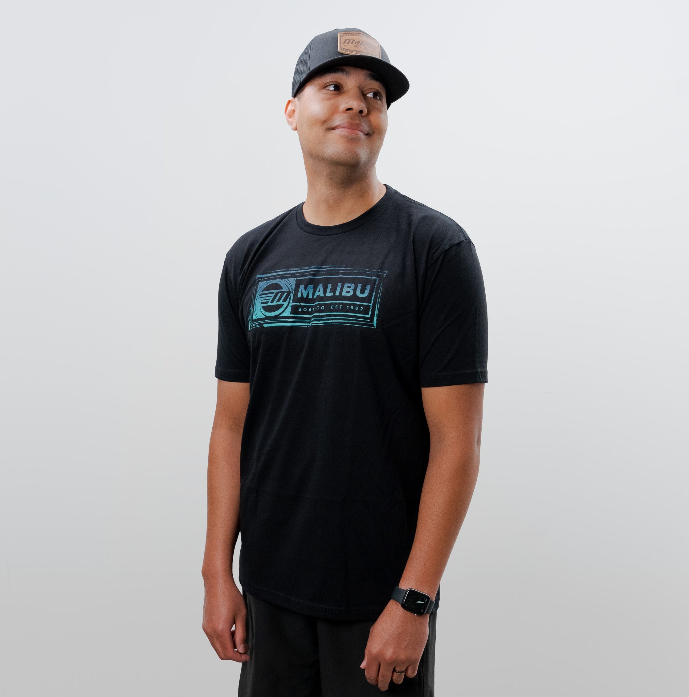 Man wearing a black Malibu Legion T-shirt, black shorts, white Nike sneakers, and a gray cap, standing against a plain white background.
