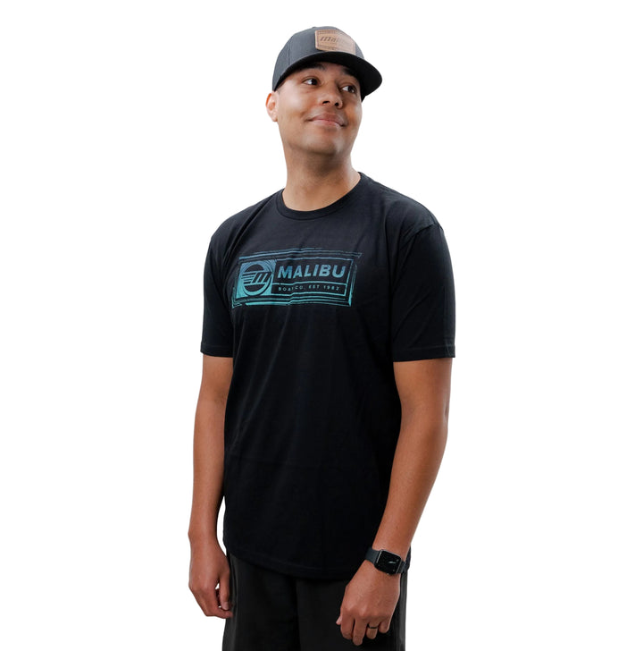 Man wearing a black Malibu Legion T-shirt, black shorts, white Nike sneakers, and a gray cap, standing against a plain white background.