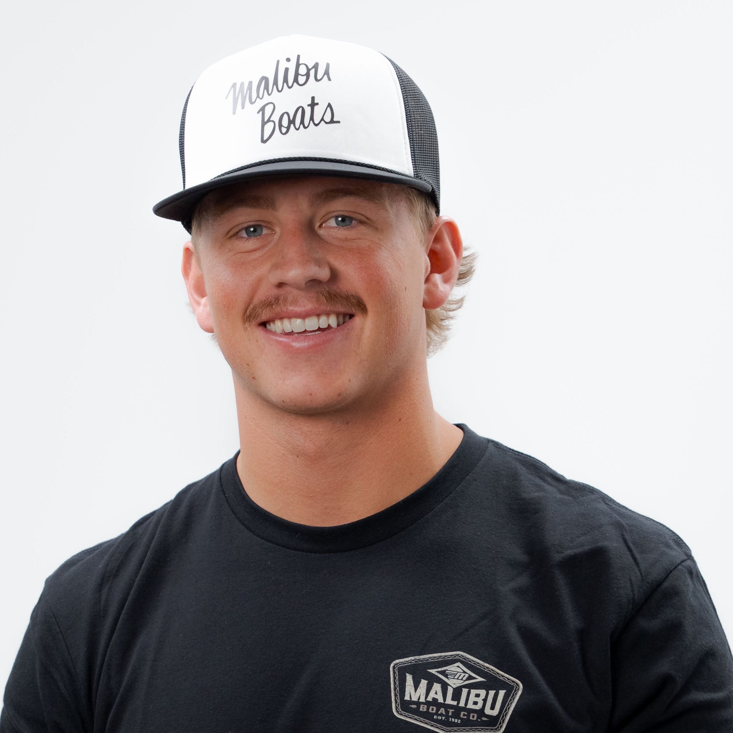 Man wearing a black Malibu Factory Trucker Hat with mesh back and front logo, smiling against a plain white background.