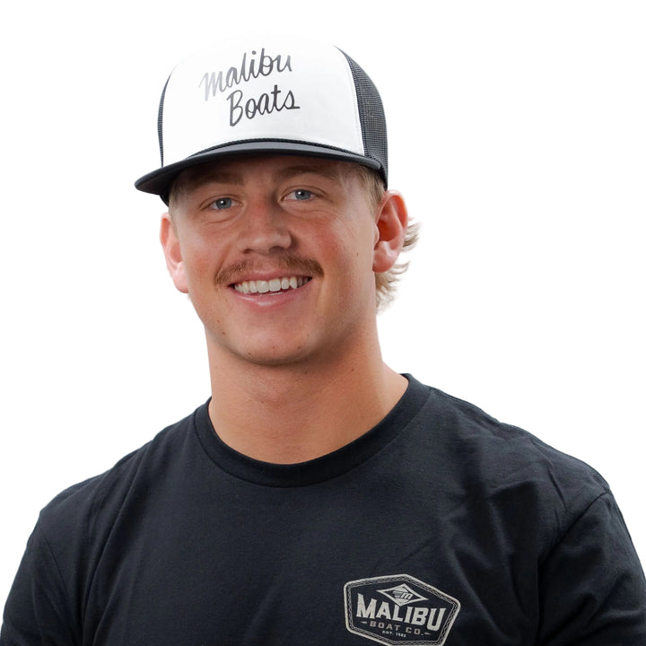 Man wearing a black Malibu Factory Trucker Hat with mesh back and front logo, smiling against a plain white background.