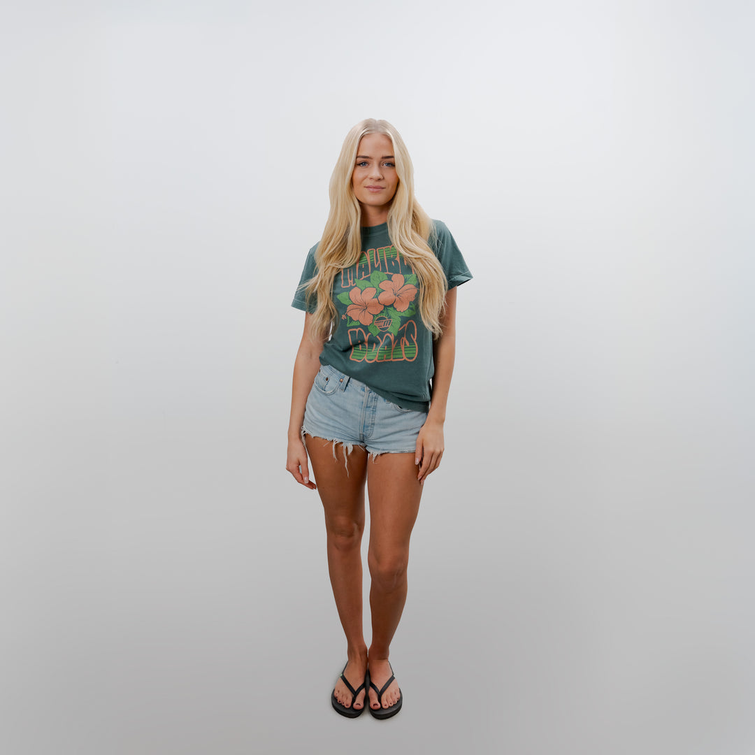 Young woman wearing a green Malibu Aloha Tee with floral graphics, paired with light denim shorts and flip-flops, standing against a plain white background.